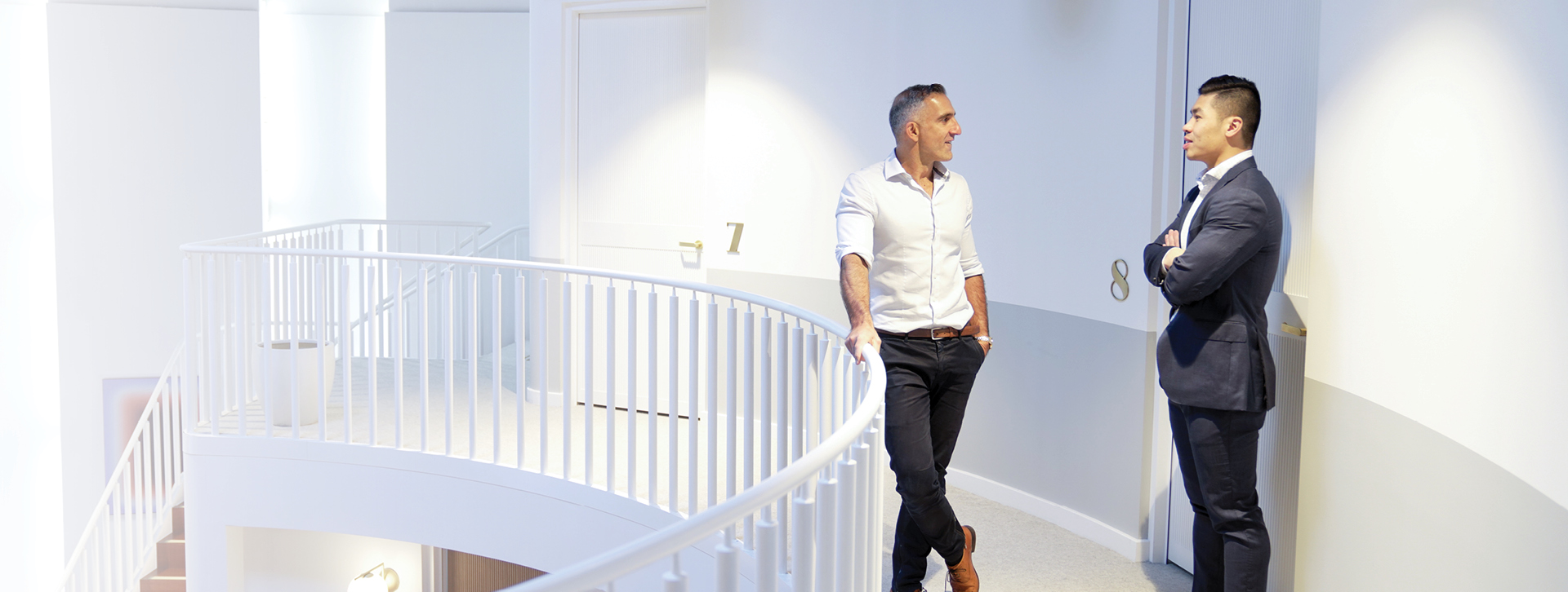 Two men in a modern, brightly lit office corridor stand near doors labeled '7' and '8.' One leans against a railing while the other stands with arms crossed, engaged in conversation. The scene feels inviting, as if encouraging us to make contact and join them.