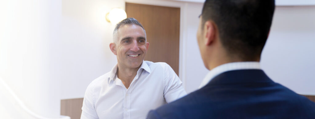 Two men, one in a white shirt and the other in a dark suit, are having a conversation in a brightly lit, modern indoor setting. Our man in the white shirt is facing the camera, smiling, while the other is seen from behind.