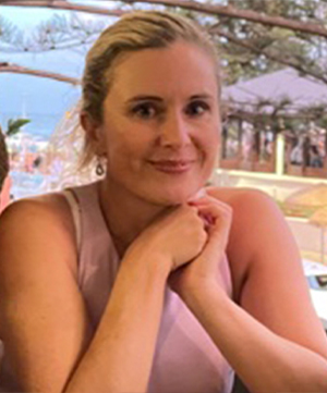 A woman with light hair and a pink sleeveless top sits at a table outdoors, resting her chin on her hands and smiling. Trees and a body of water are visible in the background, creating a serene scene perfect for contemplating life's dreams, possibly even considering car loans for her next adventure.