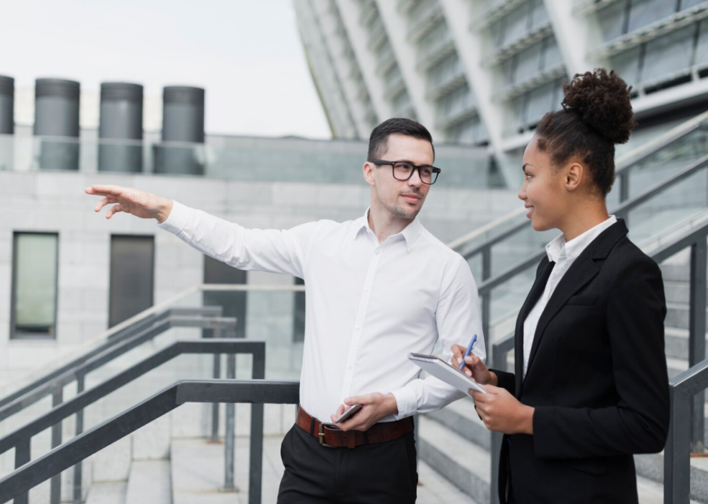 A man in a white shirt points while discussing a new program with a woman in a black blazer who is holding a notebook and pen, standing on an outdoor staircase next to a modern building.