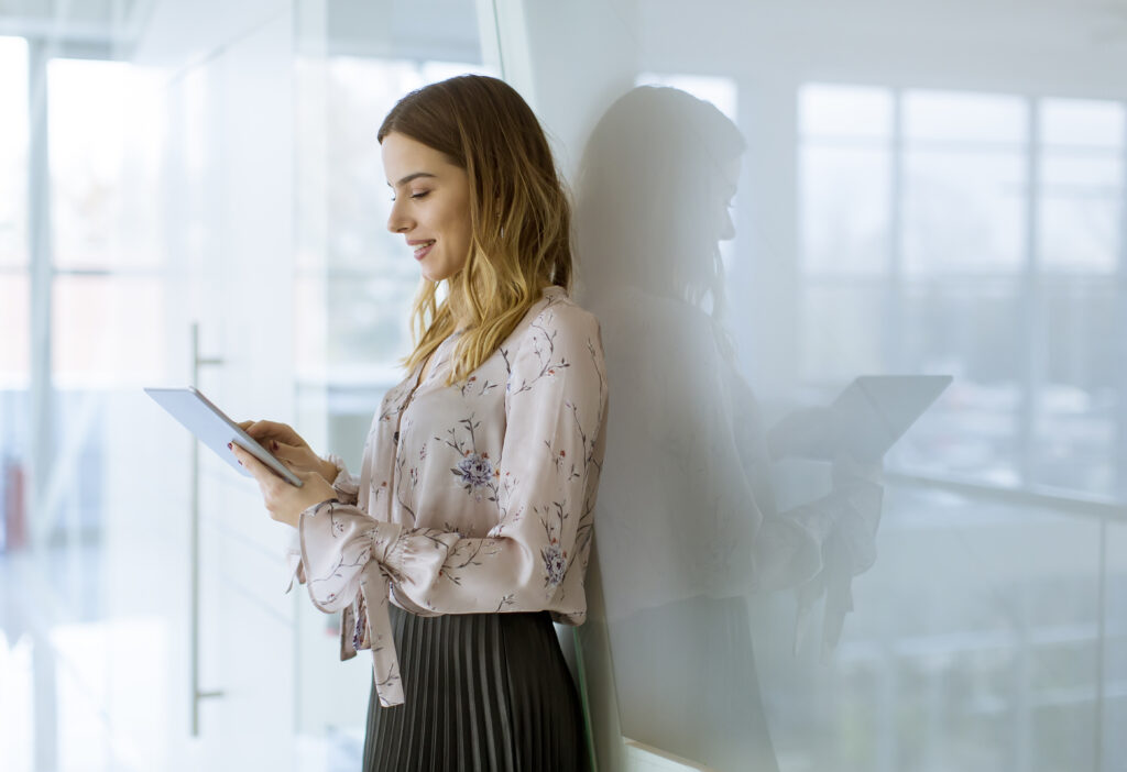 A woman stands and smiles while using a tablet in a bright, modern office setting. She leans against a glass wall, her reflection visible, as she scrolls through her company's homepage.