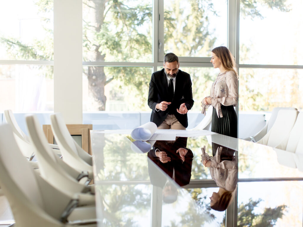 Two people in business attire stand by a large conference table with papers, engaging in a discussion. The window reveals lush greenery, adding tranquility. This setting exemplifies our difference—where every detail fosters collaboration and innovation.