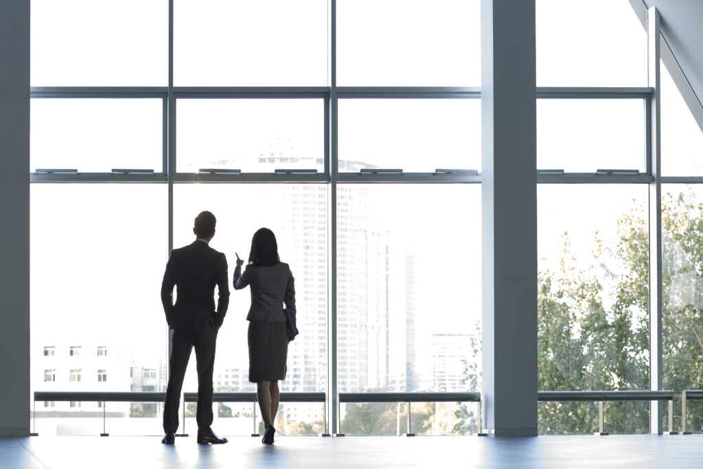 Two people in formal attire stand in front of a large window, looking out at a city skyline with tall buildings and trees, discussing finance solutions.