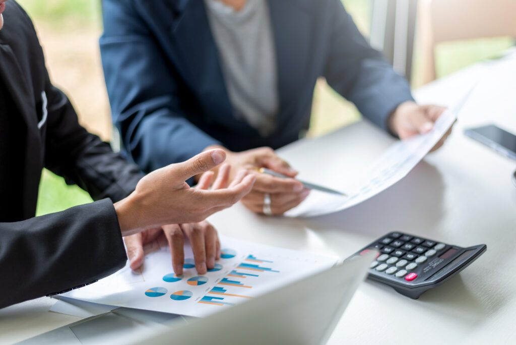 Two people in business attire sit at a table reviewing charts and graphs on paper, with a calculator and a laptop in view.