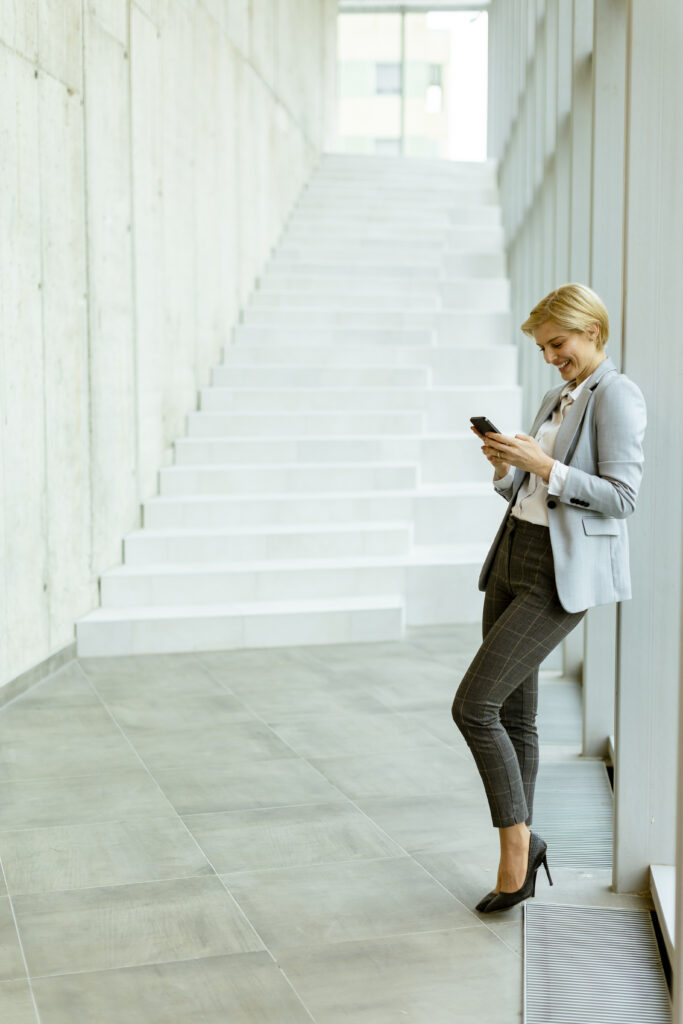 A woman in business attire stands in a modern hallway, looking at her phone and smiling as she reviews frequently asked questions. She leans against a wall near a staircase.