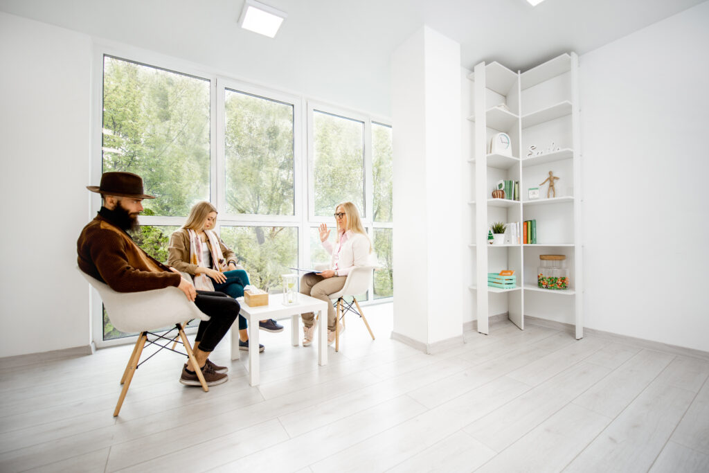 Three people sit in a well-lit, modern office with large windows; one person appears to be counseling the other two on financial practices. Shelves with books and decor are visible to the right.