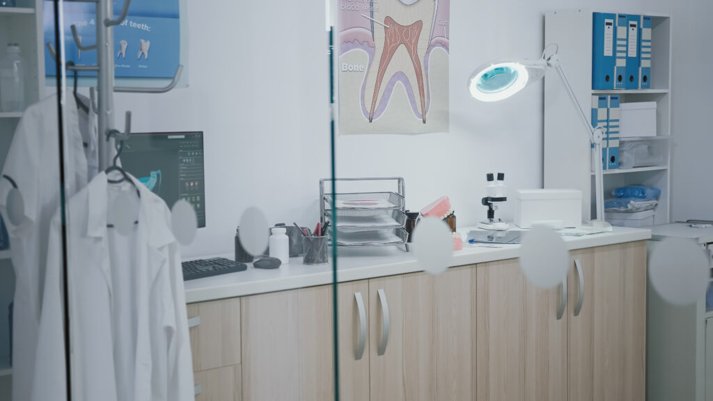 A dental office workspace, resembling a home study, featuring a computer, dental coat, microscope, dental tools, and an anatomical poster of a tooth.