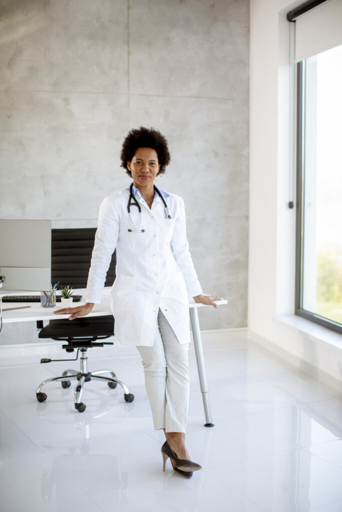 A medical professional in a white lab coat stands beside a desk in a modern office, with a stethoscope around their neck and a window in the background, ready to discuss the importance of insurance policies for comprehensive financial protection.