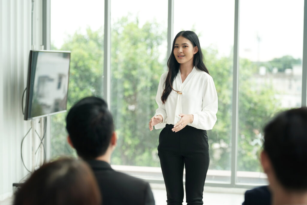 A woman in a white blouse and black pants stands in front of her team, giving a presentation in a modern office with large windows.