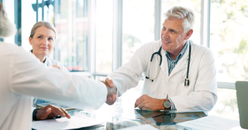 Our two doctors in white coats are seated at a conference table. One doctor on the right is shaking hands with another person outside the frame, while the other doctor watches and takes notes.