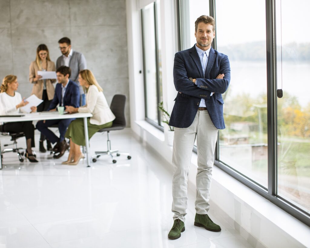 A man in a suit stands with arms crossed near a window, as a group of people conduct an About Us meeting at a table in the background of a modern office.