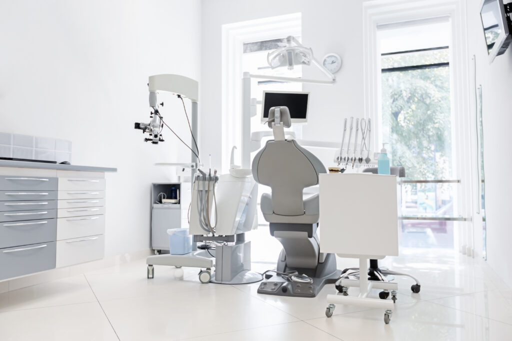 A modern dental office with a dental chair, an overhead light, a monitor, and dental instruments is impeccably organized in cabinets within a bright, white room featuring large windows. Their top-of-the-line equipment is ensured through smart equipment finance solutions.