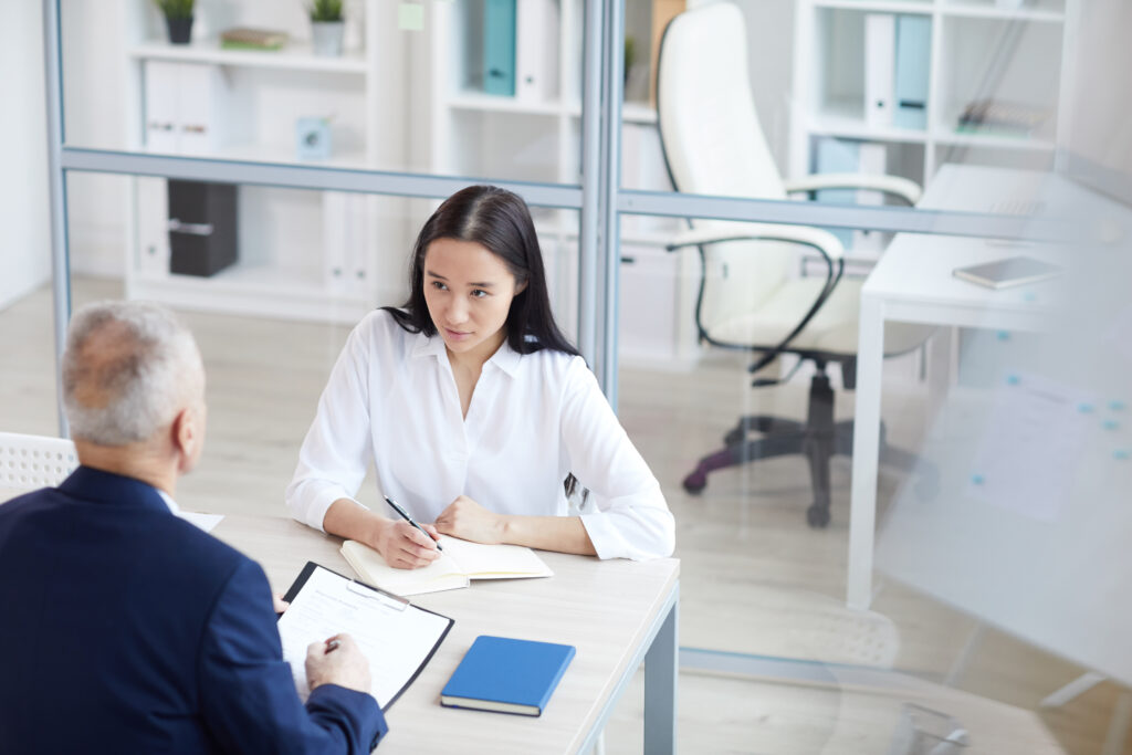 A woman in a white blouse is sitting at a desk with a notebook, engaged in a discussion with an older man in business attire. They are in a modern office, which feels almost like home, with shelves and a glass partition.