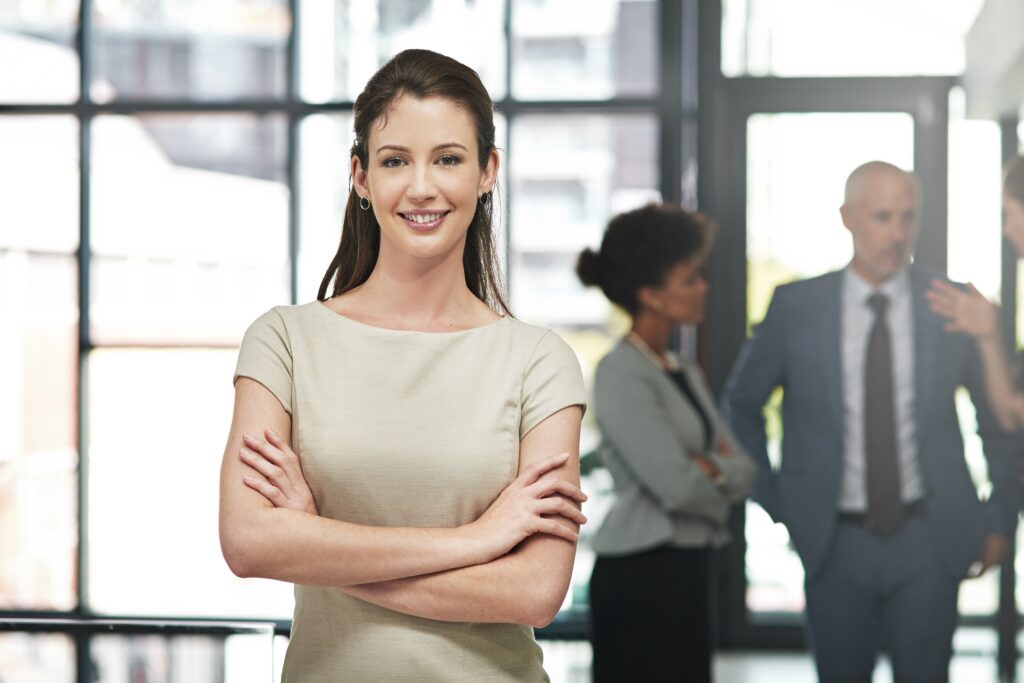 A woman with arms crossed smiles at the camera in an office setting, showcasing a positive atmosphere. Two colleagues from the team are chatting in the background.