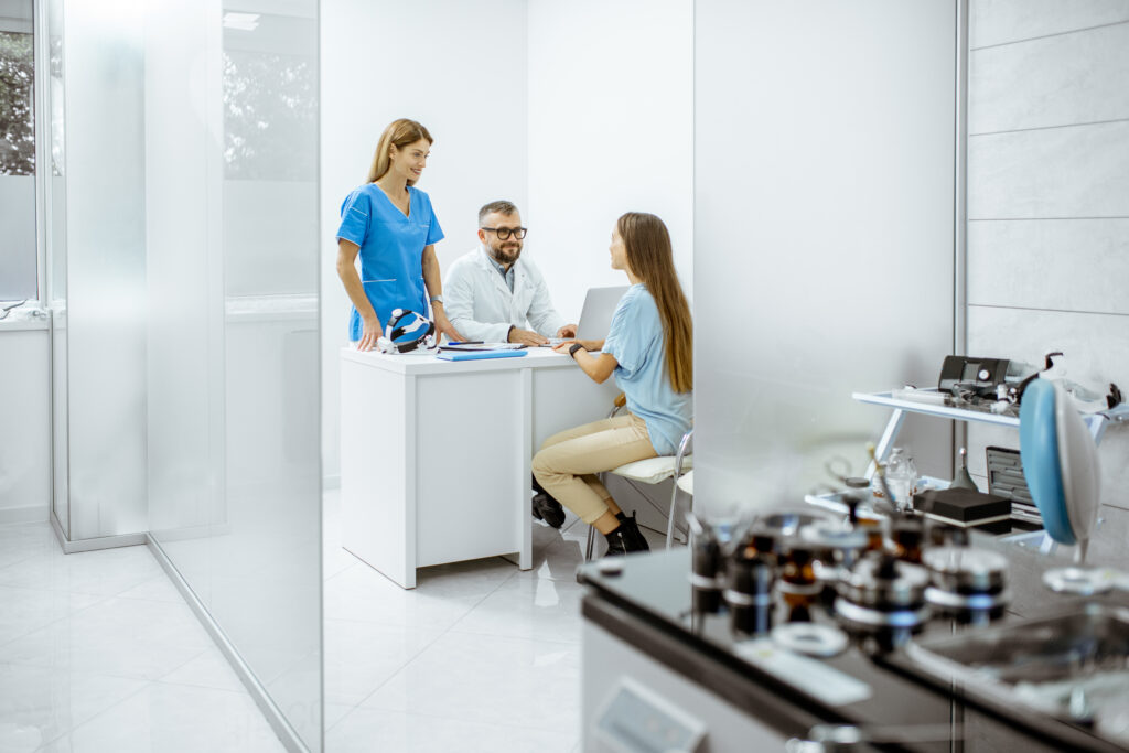 Three people are in a modern medical office. Two are seated at a desk discussing equipment finance, while a healthcare professional stands beside them. Medical equipment is visible in the foreground and background.