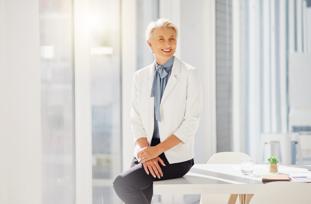 An older woman with short gray hair is sitting on the edge of a table in a brightly lit room. She is wearing a white blazer and a blue scarf, giving her the poised appearance of someone who might be discussing general insurance options with confidence and expertise.