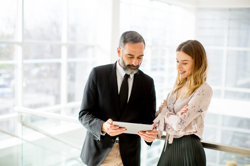 Two colleagues are standing in a bright office space, looking at a tablet together. One is a man in a suit and tie, the other is a woman in a blouse and skirt. They appear to be discussing lending practices as they review the data on the screen.