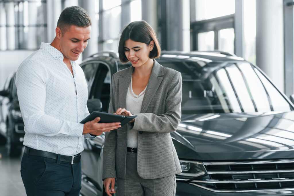 A man and a woman stand next to a black SUV in a showroom, reviewing a tablet. They're discussing car loans as they evaluate their options.