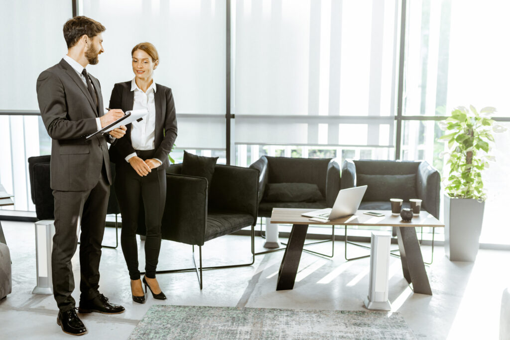 A man and woman in business attire stand in an office, engaged in a discussion. The man holds a clipboard. Office furnishings include a coffee table, laptop, and chairs where they often share ideas that highlight our difference.