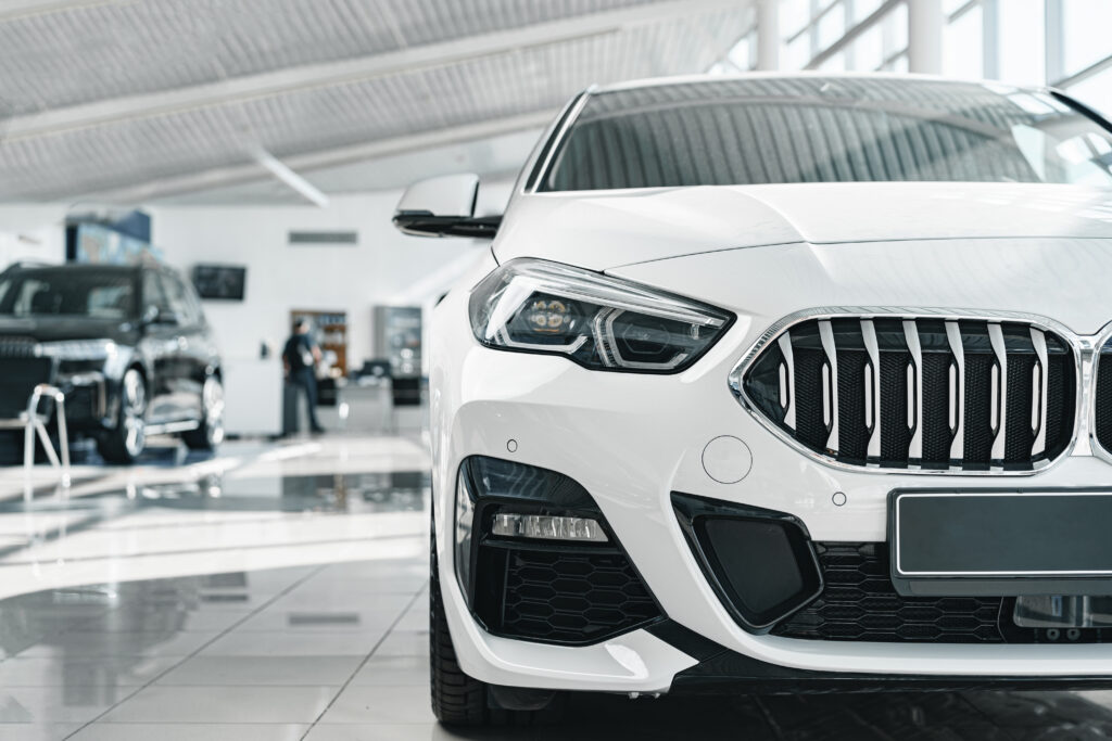 Close-up view of the front of a white car in a showroom, highlighting its sleek design, with other vehicles and a person in the background discussing car loans.