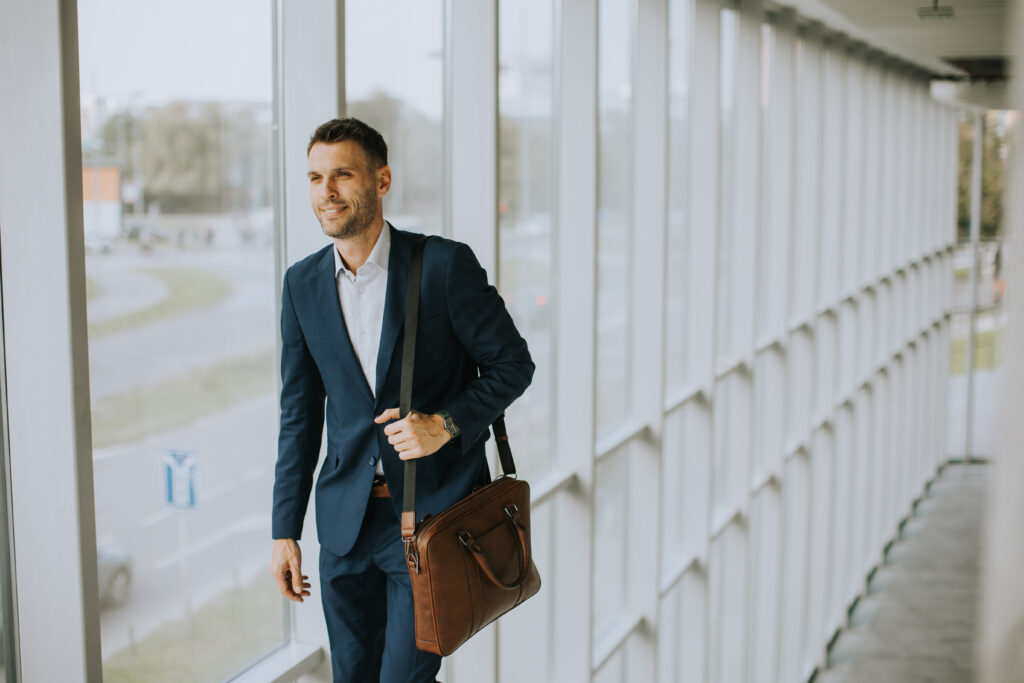 A man in a blue suit carrying a brown shoulder bag walks along a glass-walled corridor, seemingly pondering frequently asked questions.