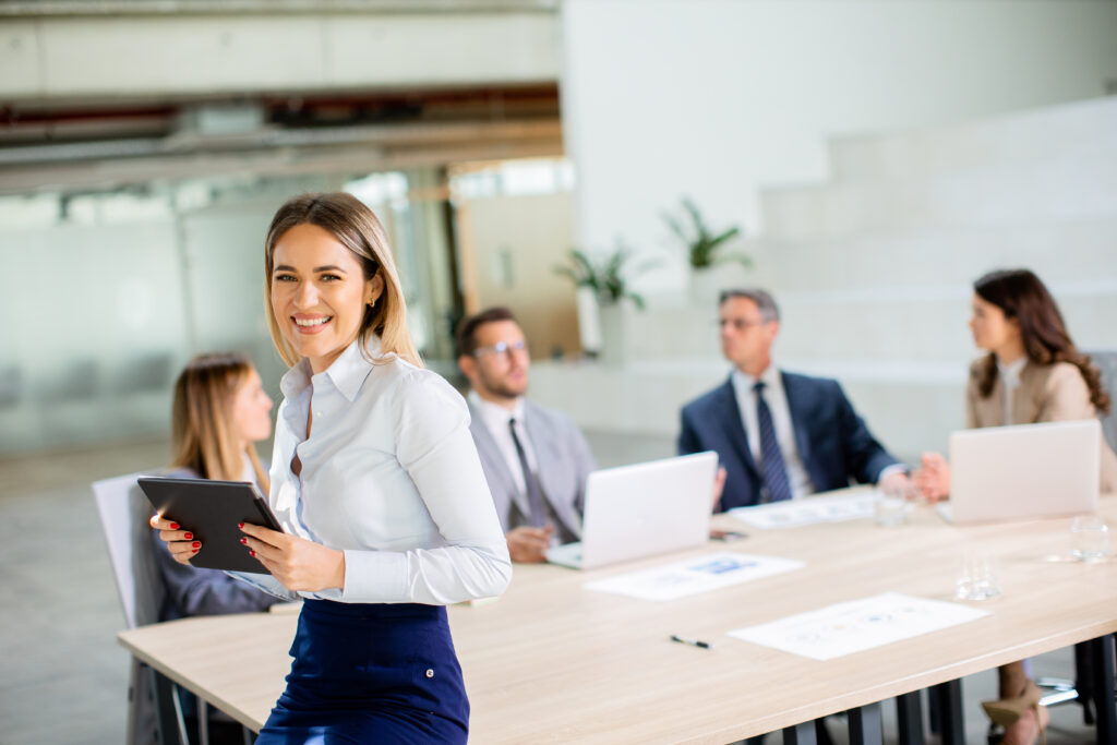 A smiling woman holding a tablet stands near a meeting table with four seated colleagues engaged in discussion about search optimization strategies.