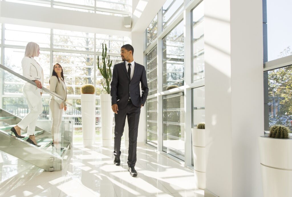 Three people, two women and one man, in professional attire discuss financing options in a modern, sunlit office atrium with large windows, glass railings, and potted cacti.