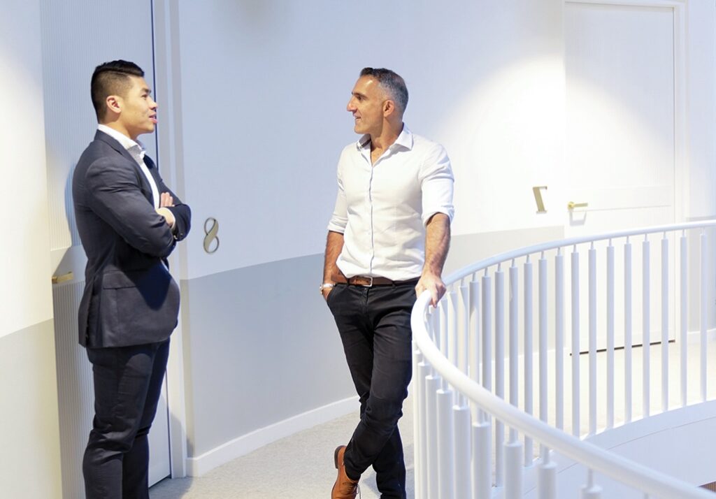 Two men are engaged in a conversation about collaboration in a brightly lit modern environment with white walls and a curved staircase.